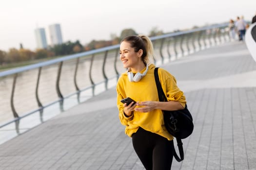 A smiling young woman walks along a riverside path in autumn, dressed in a cozy yellow sweater and headphones. The serene atmosphere captures her joyful moment.