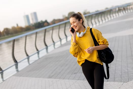 A cheerful woman dressed in a vibrant yellow sweater laughs on her phone while strolling along a picturesque riverside walkway, enjoying the warm sunshine.
