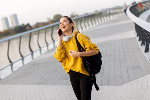 A cheerful woman dressed in a vibrant yellow sweater laughs on her phone while strolling along a picturesque riverside walkway, enjoying the warm sunshine.