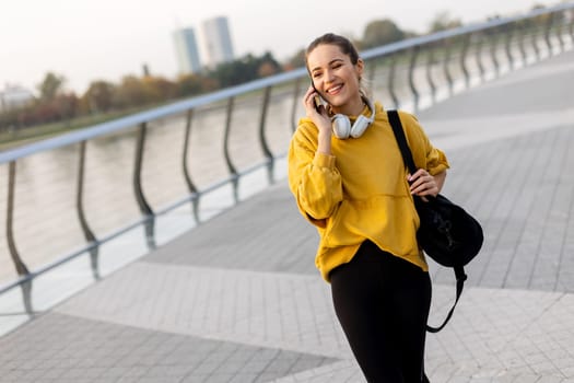 A cheerful woman dressed in a vibrant yellow sweater laughs on her phone while strolling along a picturesque riverside walkway, enjoying the warm sunshine.