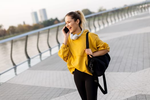 Joyful young woman strolls along a riverside walkway, engaged in a lively phone conversation while wearing a bright yellow sweatshirt and carrying a black bag
