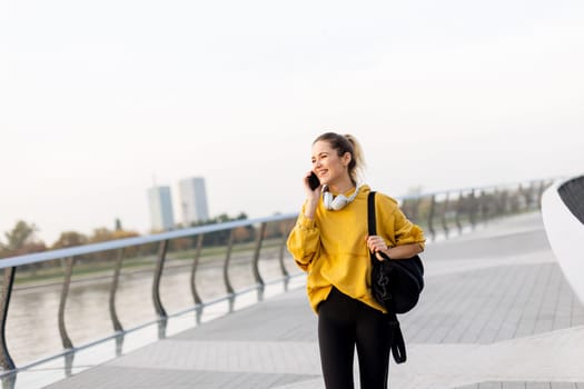 Joyful young woman strolls along a riverside walkway, engaged in a lively phone conversation while wearing a bright yellow sweatshirt and carrying a black bag