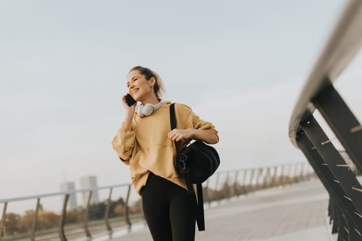 Joyful young woman strolls along a riverside walkway, engaged in a lively phone conversation while wearing a bright yellow sweatshirt and carrying a black bag