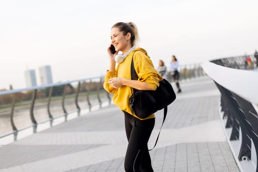 As the sun rises over the skyline, a young woman dressed in a vibrant yellow sweater walks alongside the river, captivated by her smartphone while listening to music.