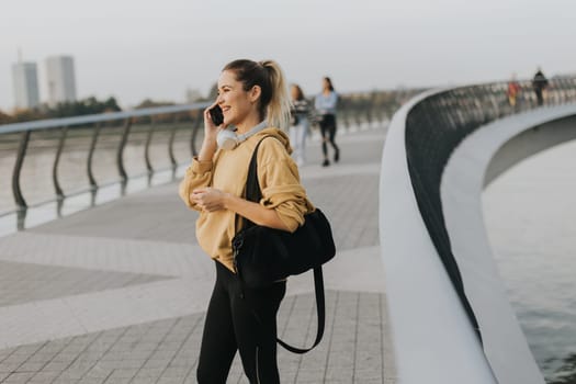 As the sun rises over the skyline, a young woman dressed in a vibrant yellow sweater walks alongside the river, captivated by her smartphone while listening to music.