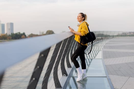 A young woman wearing a bright yellow jacket leans against a modern railing as she focuses on her phone, enjoying a tranquil riverside stroll on a pleasant afternoon.