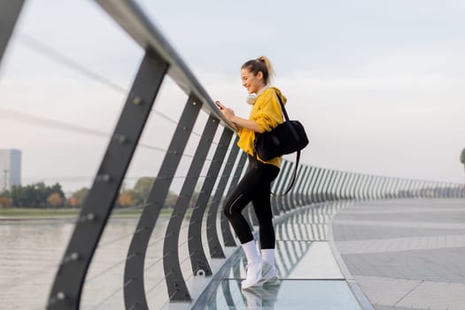A young woman wearing a bright yellow jacket leans against a modern railing as she focuses on her phone, enjoying a tranquil riverside stroll on a pleasant afternoon.