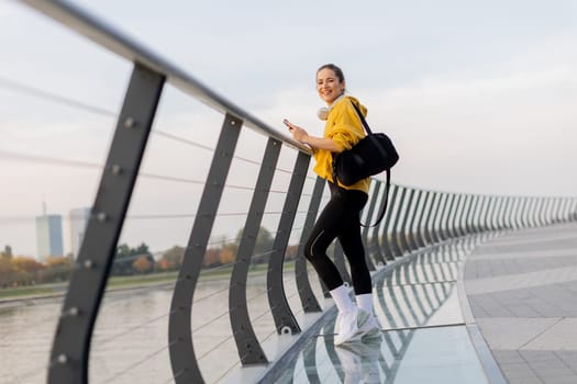 Young woman with a confident smile strolls along a sleek, glass railing by the river, dressed comfortably for an afternoon of leisure in a bright yellow hoodie and leggings.