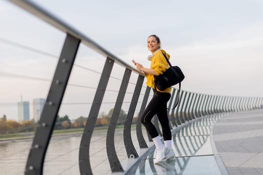 Young woman with a confident smile strolls along a sleek, glass railing by the river, dressed comfortably for an afternoon of leisure in a bright yellow hoodie and leggings.