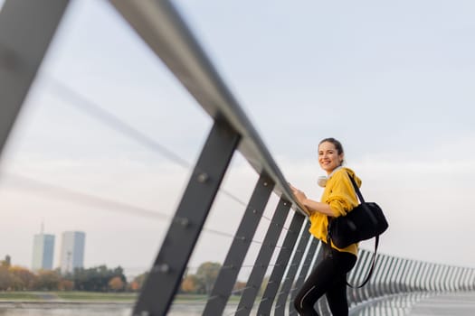 A young woman wearing a bright yellow jacket leans against a modern railing as she focuses on her phone, enjoying a tranquil riverside stroll on a pleasant afternoon.
