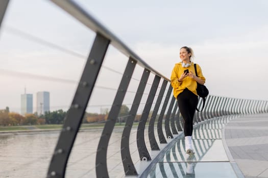 Young woman walks casually along a sleek pedestrian bridge, talking on her phone. The tranquil waters reflect a picturesque urban skyline as twilight approaches