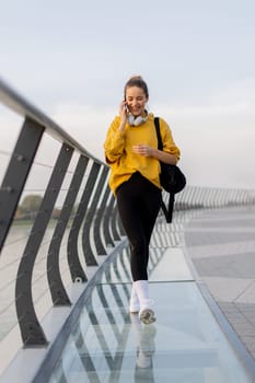 Young woman walks casually along a sleek pedestrian bridge, talking on her phone. The tranquil waters reflect a picturesque urban skyline as twilight approaches