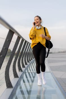 Young woman walks casually along a sleek pedestrian bridge, talking on her phone. The tranquil waters reflect a picturesque urban skyline as twilight approaches