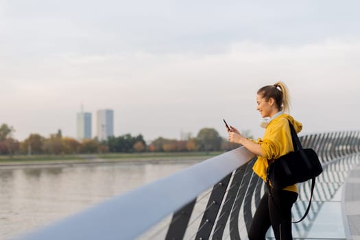 Young woman with a confident smile strolls along a sleek, glass railing by the river, dressed comfortably for an afternoon of leisure in a bright yellow hoodie and leggings.