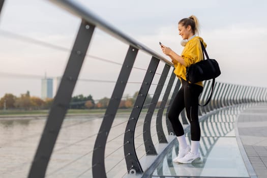 Young woman with a confident smile strolls along a sleek, glass railing by the river, dressed comfortably for an afternoon of leisure in a bright yellow hoodie and leggings.