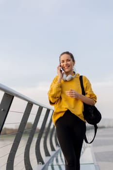 Young woman walks casually along a sleek pedestrian bridge, talking on her phone. The tranquil waters reflect a picturesque urban skyline as twilight approaches