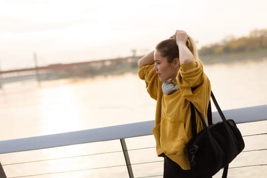 With the sun setting behind her, a cheerful young woman stands by the water, savoring a tranquil moment as she playfully pulls back her hair, feeling carefree and content