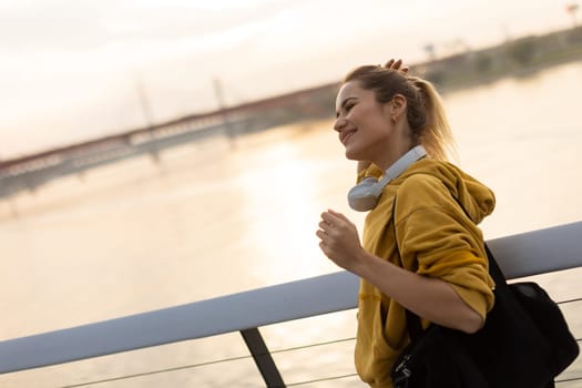 With the sun setting behind her, a cheerful young woman stands by the water, savoring a tranquil moment as she playfully pulls back her hair, feeling carefree and content