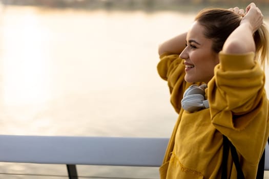 With the sun setting behind her, a cheerful young woman stands by the water, savoring a tranquil moment as she playfully pulls back her hair, feeling carefree and content