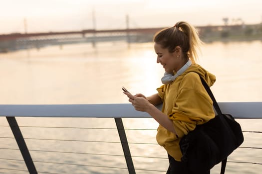 With the sun setting over the water, a young woman in a yellow hoodie stands by the riverside, absorbed in her phone while wearing headphones, embracing tranquility.