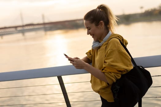 With the sun setting over the water, a young woman in a yellow hoodie stands by the riverside, absorbed in her phone while wearing headphones, embracing tranquility.