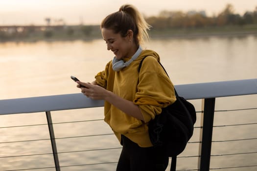 As the sun sets over the tranquil river, a young woman wearing a yellow hoodie and headphones smiles while texting, capturing a moment of joy and relaxation in nature