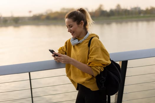 As the sun sets over the tranquil river, a young woman wearing a yellow hoodie and headphones smiles while texting, capturing a moment of joy and relaxation in nature