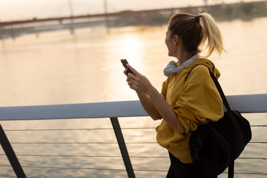 As the sun sets over the tranquil river, a young woman wearing a yellow hoodie and headphones smiles while texting, capturing a moment of joy and relaxation in nature