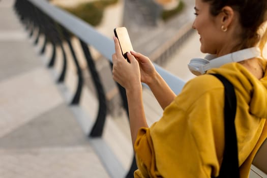 A young woman wearing a bright yellow hoodie is happily engaging with her smartphone while standing on a modern bridge. The warm sunlight enhances the vibrant atmosphere