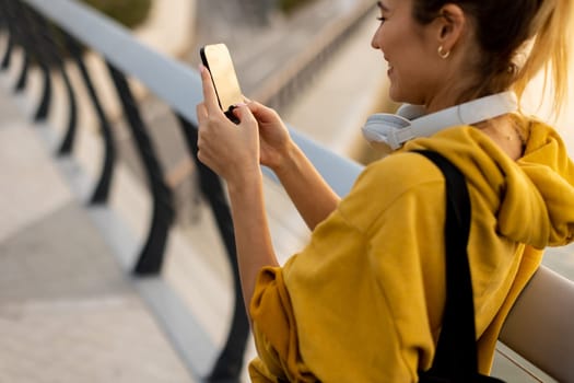 A young woman wearing a bright yellow hoodie is happily engaging with her smartphone while standing on a modern bridge. The warm sunlight enhances the vibrant atmosphere