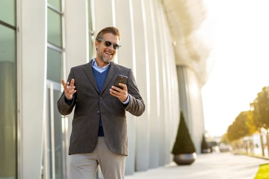 A professional man in stylish attire leisurely walks outside a sleek building, smiling as he interacts with his phone under the warm afternoon sun, exuding confidence.