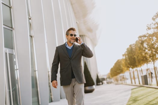 A well-dressed man strolls along a sleek pathway, chatting on his phone under the warm glow of the afternoon sun, surrounded by contemporary architecture and greenery.