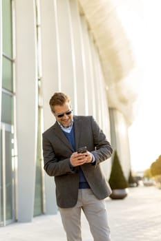 A professional man in stylish attire leisurely walks outside a sleek building, smiling as he interacts with his phone under the warm afternoon sun, exuding confidence.