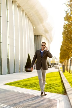 A well-dressed man strolls along a sleek pathway, chatting on his phone under the warm glow of the afternoon sun, surrounded by contemporary architecture and greenery.
