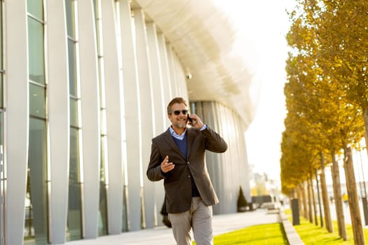 A well-dressed man strolls along a sleek pathway, chatting on his phone under the warm glow of the afternoon sun, surrounded by contemporary architecture and greenery.