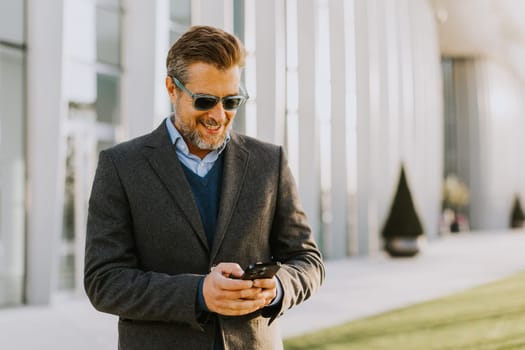 A well-dressed man stands outside a contemporary structure, smiling as he interacts with his smartphone. The bright sun casts a warm glow on his stylish outfit, reflecting happiness.