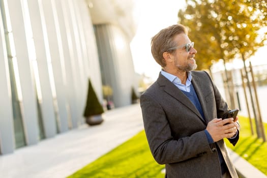 A professional man in stylish attire leisurely walks outside a sleek building, smiling as he interacts with his phone under the warm afternoon sun, exuding confidence.