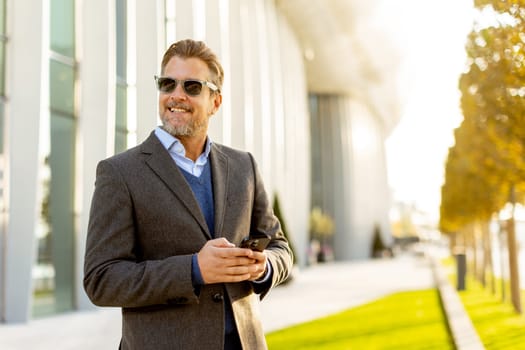 A professional man in stylish attire leisurely walks outside a sleek building, smiling as he interacts with his phone under the warm afternoon sun, exuding confidence.
