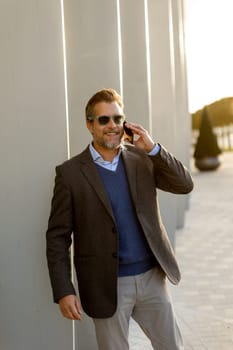 A well-dressed man is leaning casually against a sleek white wall while talking on his phone. The warm light of sunset beautifully highlights the modern architecture around him.