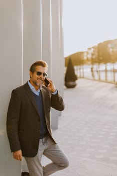 A well-dressed man is leaning casually against a sleek white wall while talking on his phone. The warm light of sunset beautifully highlights the modern architecture around him.