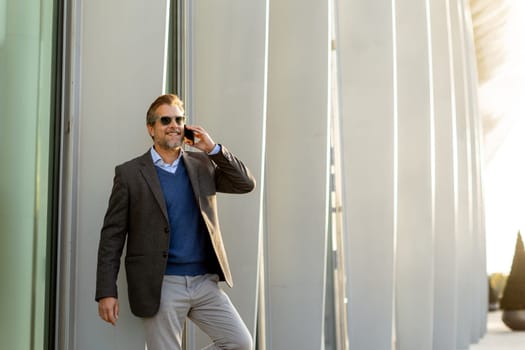 A well-dressed man is leaning casually against a sleek white wall while talking on his phone. The warm light of sunset beautifully highlights the modern architecture around him.