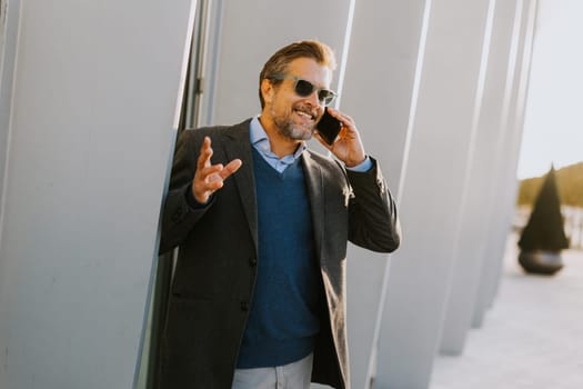 A well-dressed man enjoys a casual conversation on the phone while walking along a sunlit pathway next to contemporary buildings, creating an ambiance of sophistication.