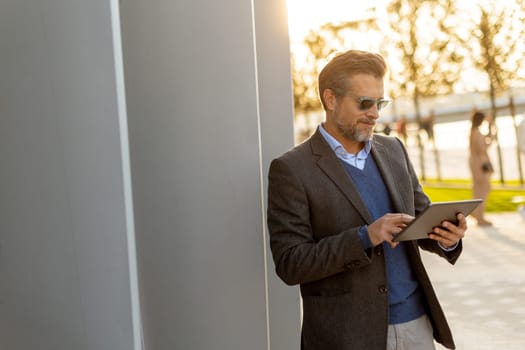 A sophisticated man stands in an urban park, engrossed in his tablet as the sun sets behind him, casting a warm golden glow over the tranquil environment.