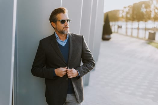 A confident man with sunglasses stands casually against a modern gray wall, savoring the warmth of a sunny day while dressed in chic, sophisticated attire.