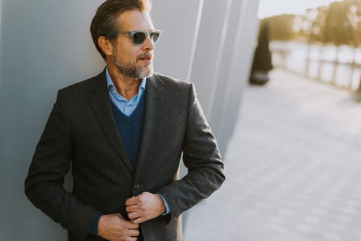 A man is seen adjusting the buttons of his blazer, embodying sophistication with sunglasses on. He stands against a modern architectural backdrop, radiating confidence in the evening glow.