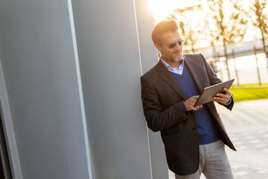 A man in a tailored suit stands confidently against a sleek wall, engrossed in his tablet as the sun illuminates the park around him, creating a joyful atmosphere.