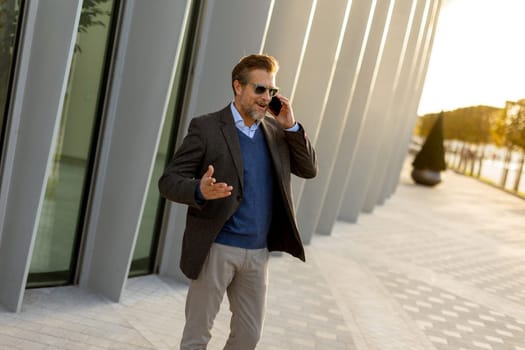 A well-dressed man enjoys a casual conversation on the phone while walking along a sunlit pathway next to contemporary buildings, creating an ambiance of sophistication.