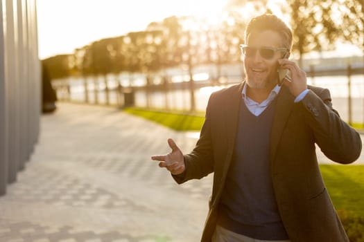 A well-dressed man enjoys a casual conversation on the phone while walking along a sunlit pathway next to contemporary buildings, creating an ambiance of sophistication.
