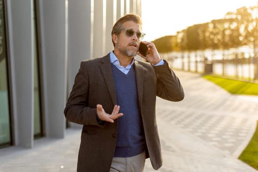 A well-dressed man enjoys a casual conversation on the phone while walking along a sunlit pathway next to contemporary buildings, creating an ambiance of sophistication.