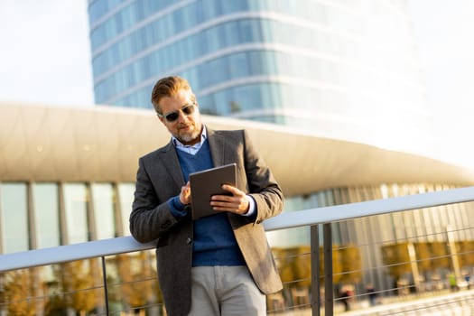 A stylish man stands on a balcony, engaging with a tablet against a backdrop of sleek architecture.